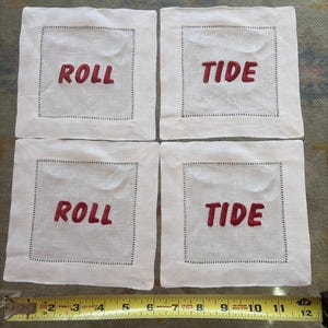 Könnte beinhalten: Vier weiße Leinenuntersetzer mit gesticktem rotem Text "ROLL TIDE". Jeder Untersetzer hat eine genähte Bordüre. Ein gelbes Maßband befindet sich im Vordergrund.