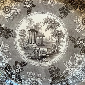 May include: A decorative plate with a detailed black and white floral and landscape design. The center features a circular scene with classical ruins, trees, and figures. The outer rim is adorned with large floral patterns.