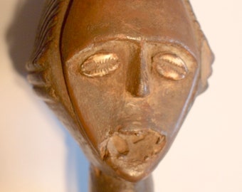 May include: A wooden sculpture of a person's head with a closed-eye expression. The sculpture is brown and has a textured surface. The head is wearing a headdress with a braided design.