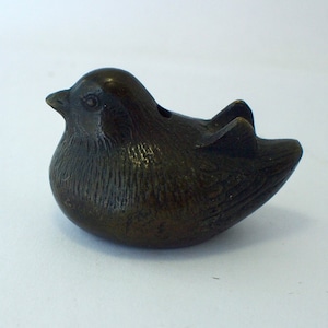 Puede incluir: Una pequeña figurita de pájaro de bronce con una superficie texturizada. El pájaro está acostado de lado con la cabeza girada hacia la izquierda.