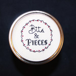 May include: A white circular fabric with a black embroidered border and red dots. The text "Bits & Pieces" is embroidered in black.