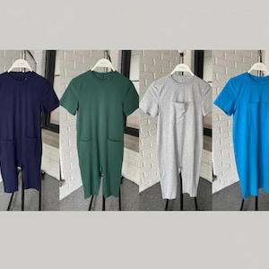 May include: Four short-sleeved jumpsuits in navy blue, green, grey, and blue. Each jumpsuit has a round neckline, a front pocket, and two side pockets. The jumpsuits are hanging on white hangers against a white brick wall.
