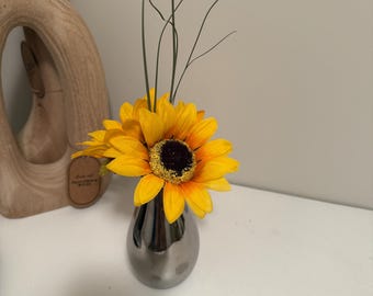 May include: A small, silver vase holds bright yellow sunflowers with dark brown centers and green stems. The vase is teardrop-shaped and sits on a white surface. A wooden sculpture is visible in the background.