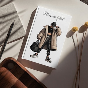Może przedstawiać: Biały planer z tekstem "Planner Girl" napisanym czarnym, ozdobnym pismem. Okładka przedstawia czarno-białą ilustrację osoby w brązowym płaszczu, czarnych spodniach i czarnym kapeluszu.