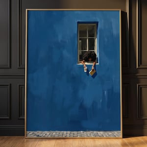 Könnte beinhalten: Ein gerahmter Wandkunstdruck zeigt eine leuchtend blaue Wand mit einem einzelnen Fenster. Eine Person mit dunklem, lockigem Haar lehnt sich aus dem Fenster und hält eine weiße Kaffeetasse sowie ein geschlossenes Buch über einem grauen Pflaster.