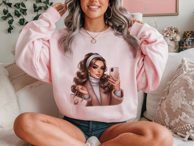 Pu&ograve; includere: Una felpa rosa con una stampa grafica di una donna con i capelli castani che indossa un cappotto marrone e tiene un telefono. La donna indossa una fascia bianca e una collana d'oro.