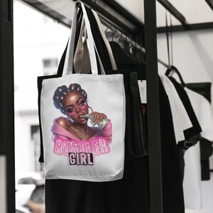 Op de afbeelding: Witte tote bag met een roze en witte illustratie van een vrouw met een zonnebril die geld in haar mond houdt. De tekst "Hustler Girl" is in roze op de tas gedrukt.