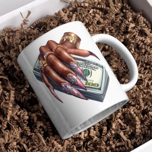 May include: White ceramic mug featuring a colourful illustration of a hand with long, glittery nails holding a stack of US dollar bills. The hand is wearing several gold rings.