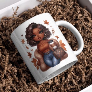 May include: White ceramic mug with a colorful illustration of a woman with brown hair and brown eyes wearing a brown sweater, blue jeans, and a fur coat. She is holding a coffee cup and a brown purse with gold accents. The mug has a brown handle and is surrounded by fall leaves.