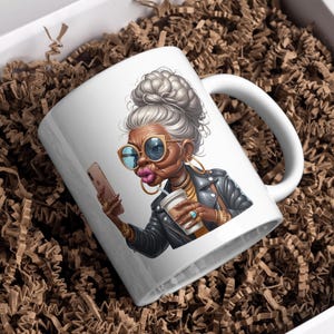 Pode incluir: Caneca de cer&acirc;mica branca com uma ilustra&ccedil;&atilde;o colorida de uma mulher com cabelo grisalho, &oacute;culos de sol e uma jaqueta de couro. Ela est&aacute; segurando um smartphone em uma m&atilde;o e uma x&iacute;cara de caf&eacute; na outra.
