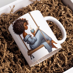 May include: White ceramic mug with a graphic of a woman with brown curly hair sitting on a swing and reading a book while holding a cup of coffee. The woman is wearing a blue robe.