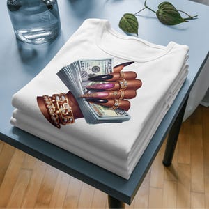 May include: White t-shirt featuring a graphic of a hand holding a stack of US dollar bills. The hand is adorned with gold rings and has long, manicured nails.