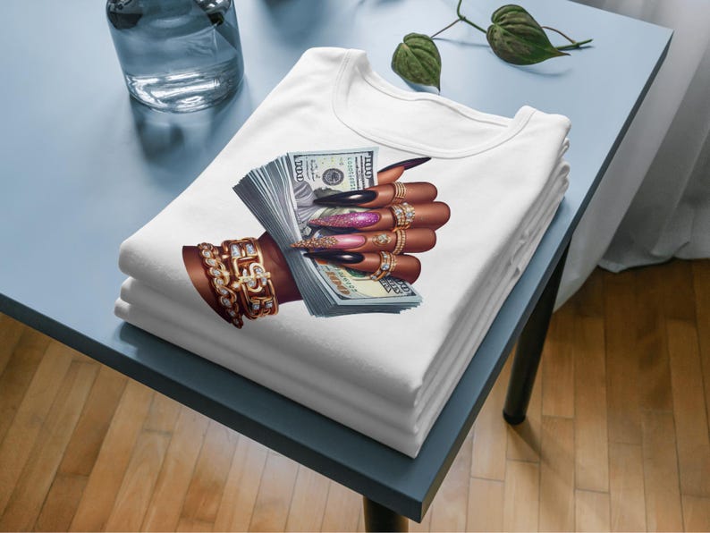May include: White t-shirt featuring a graphic of a hand holding a stack of US dollar bills. The hand is adorned with gold rings and has long, manicured nails.