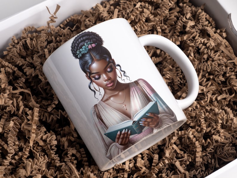 May include: White ceramic mug with a colorful illustration of a woman with dark skin reading a book. She has a floral hairpiece and is wearing a light pink and blue dress.
