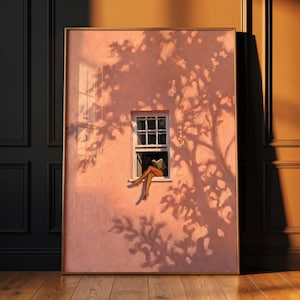 May include: A framed rectangular wall art print featuring a textured pink wall with a central white window. A pair of legs extends from the window sill, and the scene is overlaid with the dark, intricate shadow of a tree, creating a high-contrast effect.