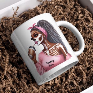 May include: White ceramic coffee mug with a pink and black illustration of a woman with a skeleton design and the text "Spooky Mom".