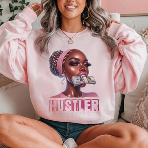 Op de afbeelding: Roze sweatshirt met een afbeelding van een vrouw met een stapel geld in haar mond. De tekst "Hustler" is onder de afbeelding gedrukt.