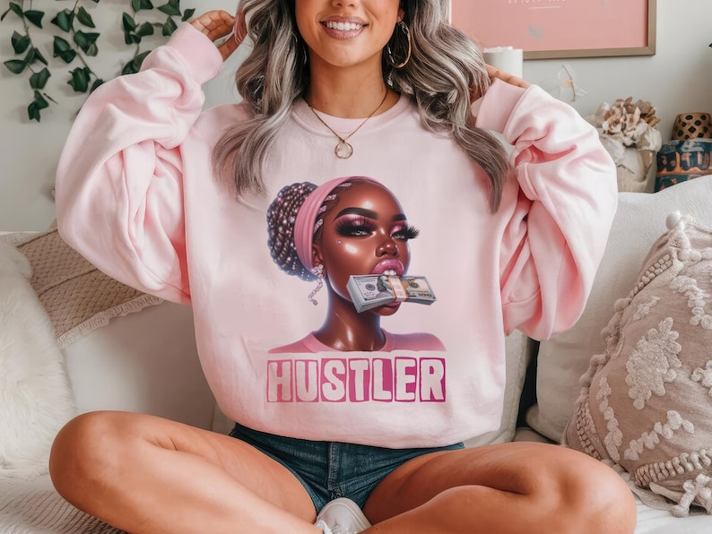 Op de afbeelding: Roze sweatshirt met een afbeelding van een vrouw met een stapel geld in haar mond. De tekst "Hustler" is onder de afbeelding gedrukt.