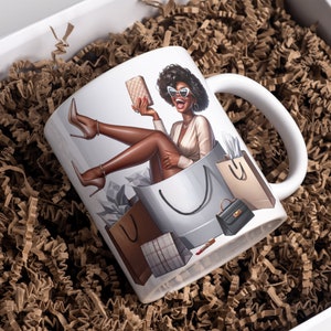 May include: White ceramic mug with a colorful illustration of a woman sitting in a shopping bag with multiple shopping bags around her. The woman is wearing sunglasses and a white top.