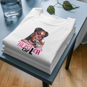 Op de afbeelding: Wit T-shirt met een afbeelding van een vrouw die een roze zonnebril draagt en een stapel geld bijt. De tekst "Hustler Girl" is in roze en zwarte letters op het shirt gedrukt.