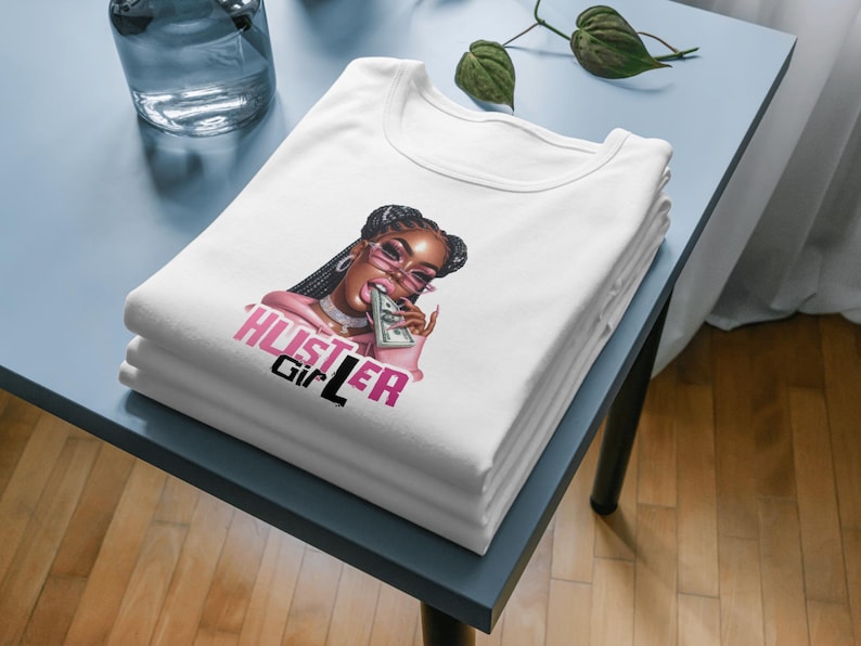 Op de afbeelding: Wit T-shirt met een afbeelding van een vrouw die een roze zonnebril draagt en een stapel geld bijt. De tekst "Hustler Girl" is in roze en zwarte letters op het shirt gedrukt.