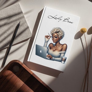 May include: A white notebook with the text "Lady Boss" in black cursive script. The notebook features an illustration of a woman with blonde hair wearing glasses and a white sweater, sitting at a desk with a laptop and a cup of coffee.
