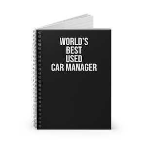 以下が含まれることがあります： 表紙に「WORLD'S BEST USED CAR MANAGER」という白い文字が書かれた黒いスパイラルノート。ノートは少し開いており、内側の白い罫線入りのページが見え、左側には黒い金属製のスパイラル綴じが施されています。