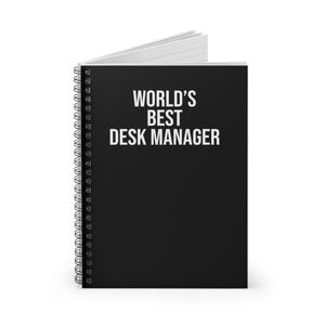 以下が含まれることがあります： 黒いスパイラル綴じのノートで、表紙には白い文字で「WORLD'S BEST DESK MANAGER」と印刷されています。ノートは少し開いており、黒い表紙の内側にある白い罫線入りのページと金属製のワイヤー綴じが見えています。