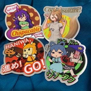 Op de afbeelding: Vier zilveren stickers met anime-personages. De eerste sticker toont een meisje met paars haar dat een groen shirt draagt en de tekst "Are you Chupacable or nah". De tweede sticker toont een meisje met bruin haar dat een wit shirt draagt en de tekst "Code Consultant". De derde sticker toont een meisje met geel haar dat een gele en oranje jurk draagt en de tekst "Haniwa,  Go!" De vierde sticker toont twee meisjes, een met blauw haar en een met zwart haar, die kleurrijke jurken dragen en de tekst "Doki Doki".
