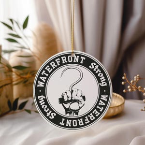 May include: Black and white round ceramic ornament with the text "Waterfront Strong" written in a circle around a hook and fist graphic.