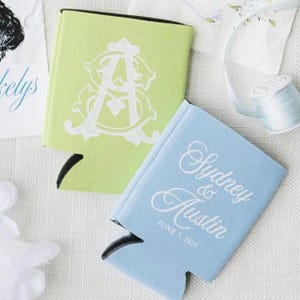 May include: Two beverage insulators, one light green with a white monogram and one light blue with the names "Sydney & Austin" and the date "June 7, 2025" in white script. The insulators are on a white surface.