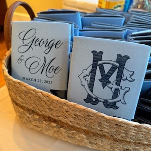 May include: A woven basket filled with light blue can coolers. One cooler displays the names "George & Moe" and the date "March 22, 2025" in black script. Another cooler features a black monogram design.