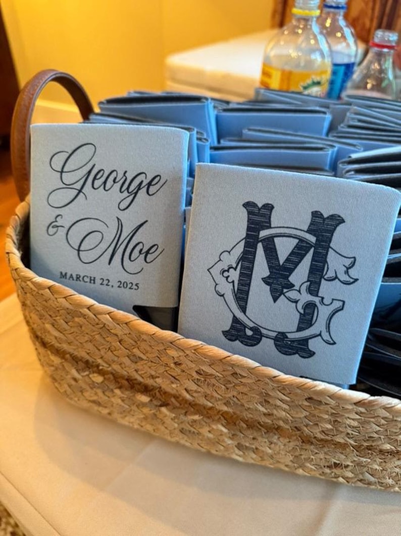 May include: A woven basket filled with light blue can coolers. One cooler has the names "George & Moe" and the date "March 22, 2025" printed in black script. Another cooler displays a black monogram.