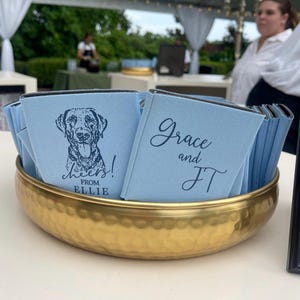 May include: A gold hammered metal bowl filled with light blue drink koozies. Some koozies have a dog illustration and the text "Cheers! From Ellie". Other koozies have the text "Grace and JT". The bowl sits on a white surface.