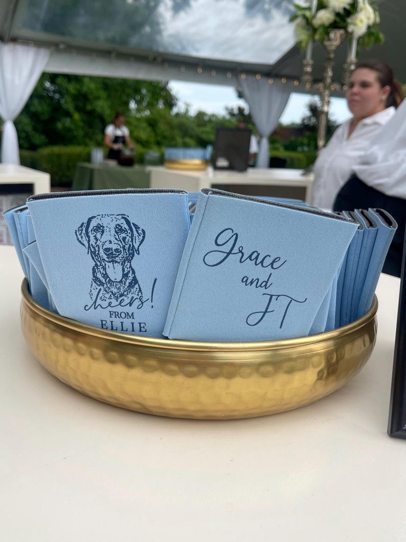May include: A gold hammered metal bowl filled with light blue drink koozies. Some koozies have a dog illustration and the text "Cheers! From Ellie". Other koozies have the text "Grace and JT". The bowl sits on a white surface.