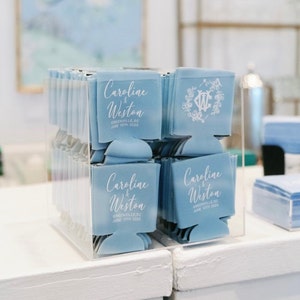 May include: A display of light blue can coolers with white text. The text reads "Caroline & Weston, Greenville, SC, June 10th 2023." Some coolers feature a floral monogram. The coolers are in a clear acrylic box.