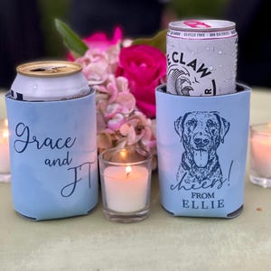 May include: Two light blue can coolers with text and graphics. One cooler has the names "Grace and JT" in script. The other has a dog graphic and the words "cheers! from ELLIE". A lit candle and flowers are in the background.