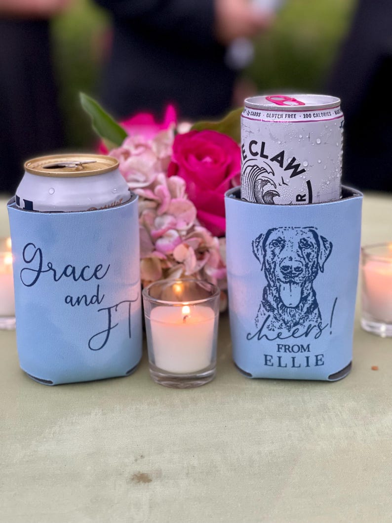 May include: Two light blue can coolers with text and graphics. One cooler has the names "Grace and JT" in script. The other has a dog graphic and the words "cheers! from ELLIE". A lit candle and flowers are in the background.