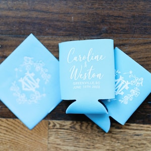 May include: Light blue wedding favors, including two square coasters and a beverage insulator. The insulator has the names "Caroline & Weston", the city, and the date "June 10th 2023" printed in white. The coasters feature a white floral design.