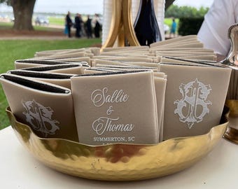 Op de afbeelding: Een verzameling beige servetten met zwarte randen, gepresenteerd in een gouden, gehamerde metalen kom. Elke servet is bedrukt met een wit monogram en de namen "Sallie & Thomas" en de locatie "Summerton, SC".