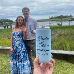 May include: A light blue can cooler with the text "Rachel & Collins Shellmore" printed on it. The cooler is decorated with a floral design and two small hearts.