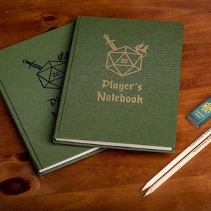Op de afbeelding: Twee groene notitieboekjes met een witte rand en een gouden inscriptie die "Player's Notebook" luidt. De notitieboekomslagen tonen een zwart-witte illustratie van een twintigzijdige dobbelsteen met een zwaard en een draak.