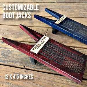 May include: Two wooden boot jacks with a distressed wood finish. The red boot jack has a wooden plaque with the text "KRUM, TEXAS - BOOTS OFF" and an American flag design. The blue boot jack has a wooden plaque with the text "GORDON BAY". Both boot jacks are 12 x 4.5 inches.