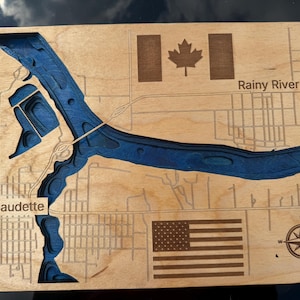 May include: A rectangular wooden map featuring a deep blue laser-cut river channel winding through the landscape. The map includes the engraved text Baudette and Rainy River, a Canadian flag, an American flag, and a compass rose in the corner.