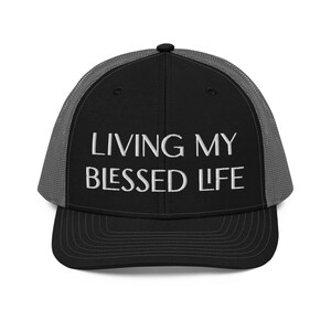 Puede incluir: Gorra de camionero negra con la parte trasera de malla gris y visera curva. La parte delantera de la gorra presenta el texto bordado "LIVING MY BLESSED LIFE" en blanco.