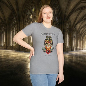 May include: A gray t-shirt with a graphic of a woman with candles on her head and the text "SAINT LUCY" and "PLAYFUL IS" printed on the front.