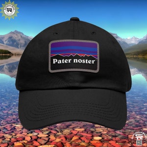 May include: Black baseball cap with a rectangular patch featuring a mountain range design in red, blue, and purple, with the text "Pater noster" in white. The cap has a curved brim and a structured crown.