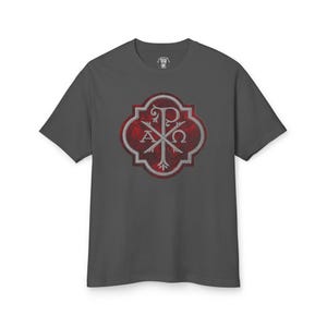 Op de afbeelding: Een donkergrijs T-shirt met een rode en zilveren afbeelding van een christelijk symbool, de Chi Rho, met de letters "P", "X", "A" en "Ω" in een decoratief kader.