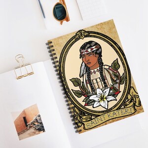 May include: A spiral bound notebook with a vintage-style illustration of a Native American woman wearing a headdress and traditional clothing. The illustration is surrounded by a gold frame and the text "SAINT KATERI" is written in a decorative font.