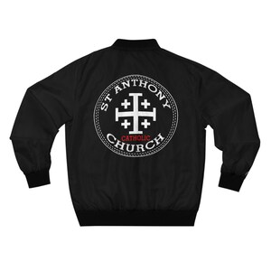 Może przedstawiać: Czarna kurtka bomberowa z białym haftowanym kołem z tyłu. Koło zawiera napis "St. Anthony Catholic Church" i biały krzyż z czterema ramionami.